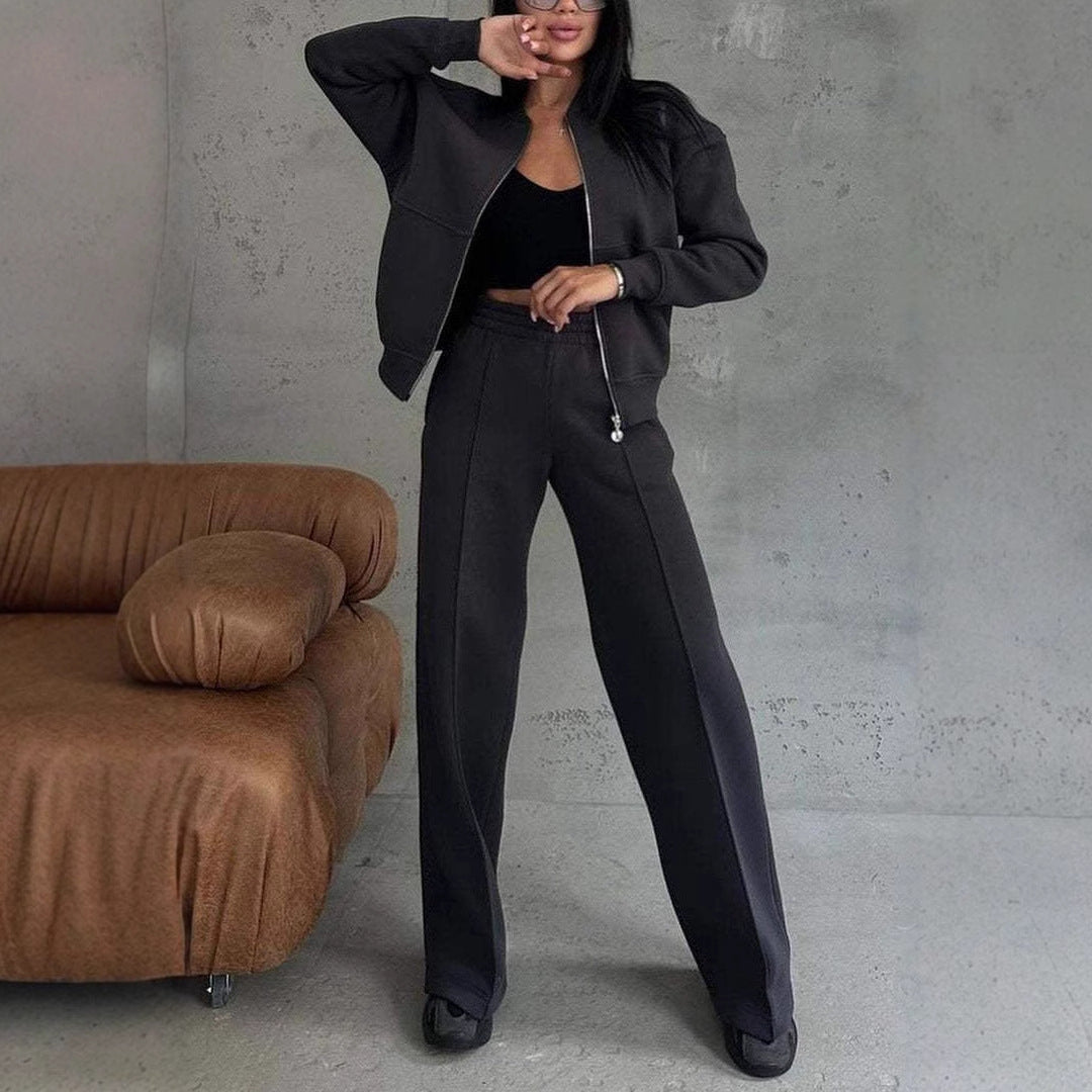 Femme en tenue décontractée chic, portant un ensemble noir avec veste et pantalon larges, posant devant un canapé marron sur fond de mur gris.