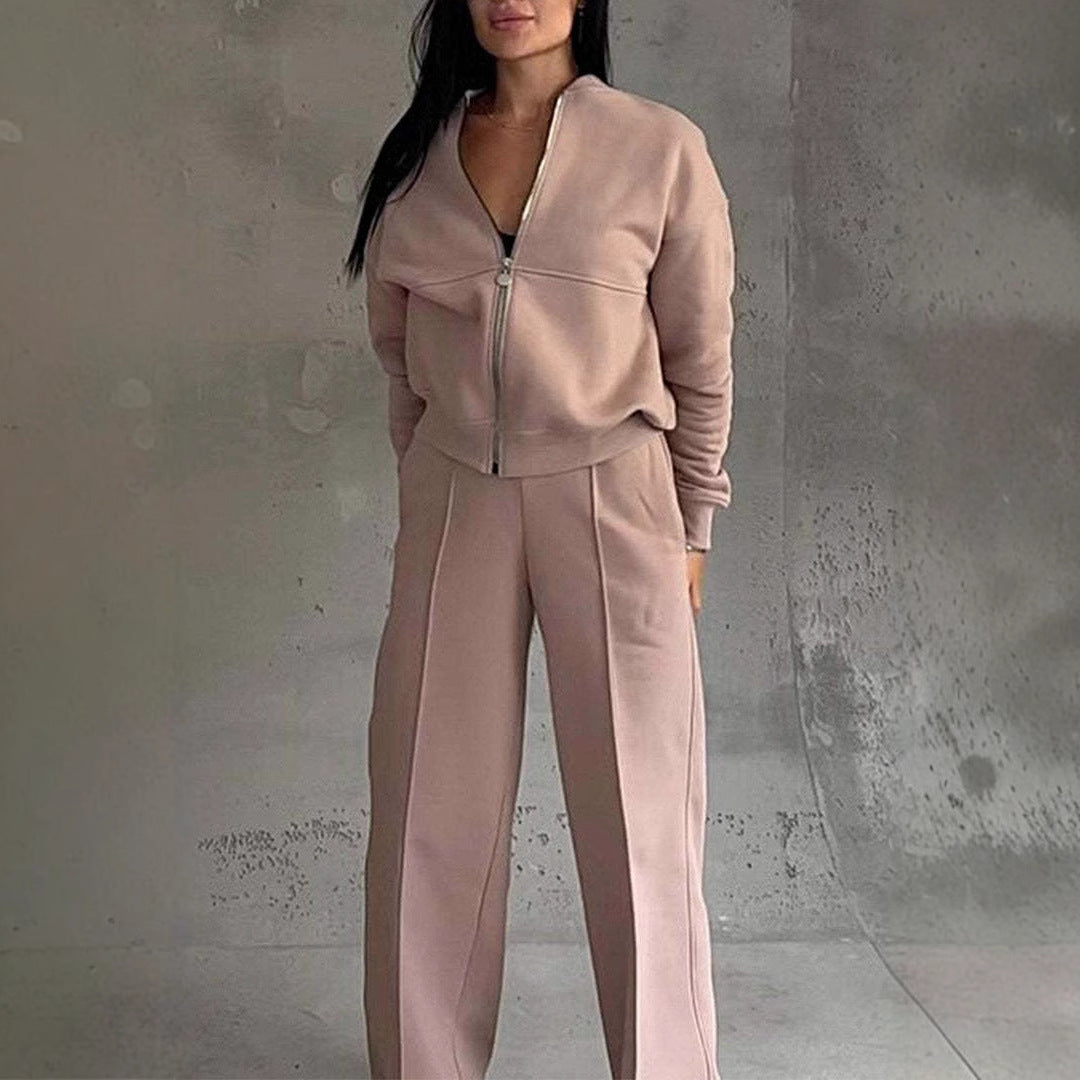 Femme portant un ensemble de survêtement beige avec veste zippée et pantalon ample, posant devant un mur gris texturé. Mode décontractée élégante.