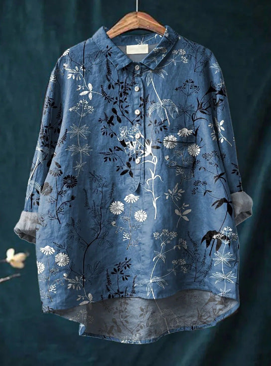 Chemise bleue à motifs floraux blancs et noirs, manches longues retroussées, sur cintre en bois, fond vert foncé. Mode élégante et décontractée.