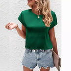 Femme en t-shirt vert et short en jean, tenant un sac à main beige, posant devant un mur blanc. Mode estivale, tenue décontractée tendance.