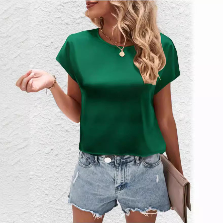 Femme en t-shirt vert et short en jean, tenant un sac à main beige, posant devant un mur blanc. Mode estivale, tenue décontractée tendance.