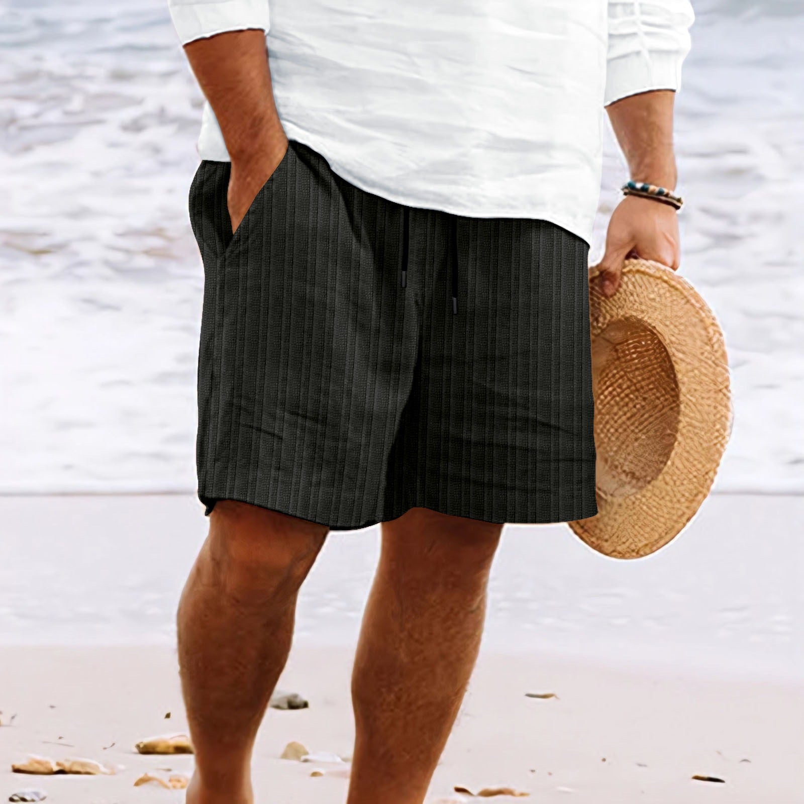 Homme en short rayé noir et chemise blanche tenant un chapeau de paille sur une plage. Mode estivale, tenue décontractée, bord de mer.