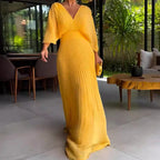 Robe longue jaune plissée élégante, femme debout dans un salon moderne avec mobilier en bois, lumière naturelle, ambiance tropicale.