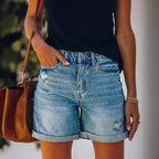 Femme en short en jean décontracté, main dans la poche, portant un sac en cuir marron et un haut noir. Mode estivale, style décontracté.