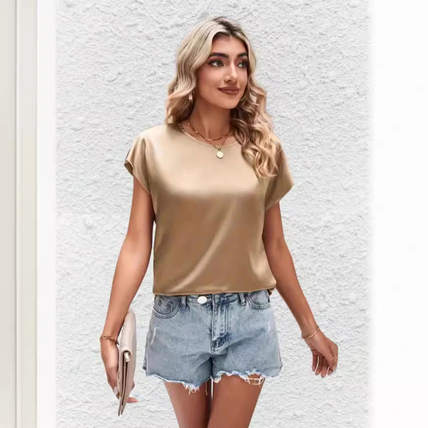 Femme blonde portant un t-shirt beige et un short en jean déchiré, tenant une pochette beige, posant devant un mur blanc texturé. Mode estivale décontractée.