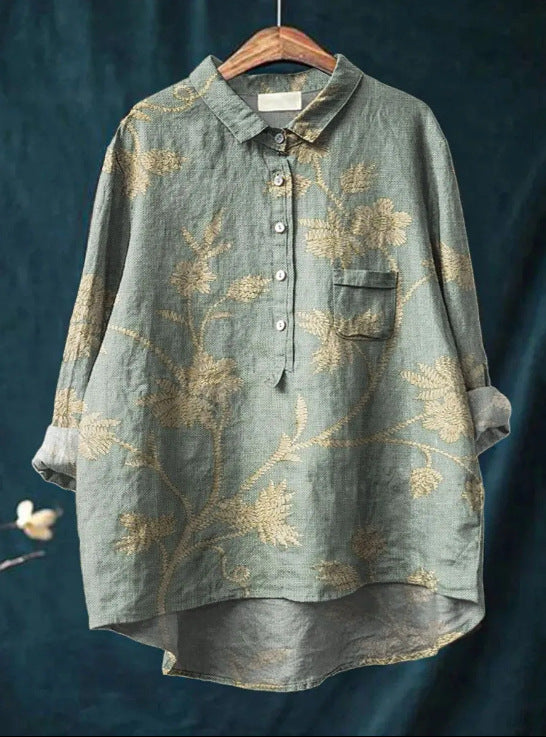 Chemise en lin vert avec motifs floraux dorés, manches longues retroussées, col classique, poche poitrine, suspendue sur un cintre en bois.