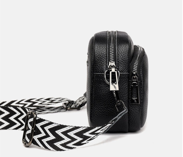 Sac bandoulière en cuir noir avec fermeture éclair argentée et sangle à motif zigzag noir et blanc, design moderne et élégant.
