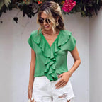 Femme portant un chemisier vert à volants et un short blanc déchiré, lunettes de soleil, posant devant un mur blanc avec fleurs. Mode estivale chic.