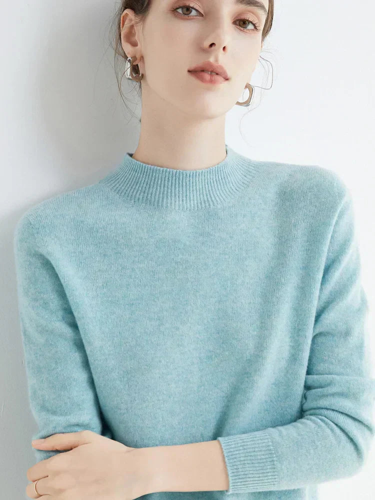 Femme portant un pull en cachemire bleu clair, col rond, manches longues, posant contre un mur blanc. Mode élégante, tendance automne-hiver.