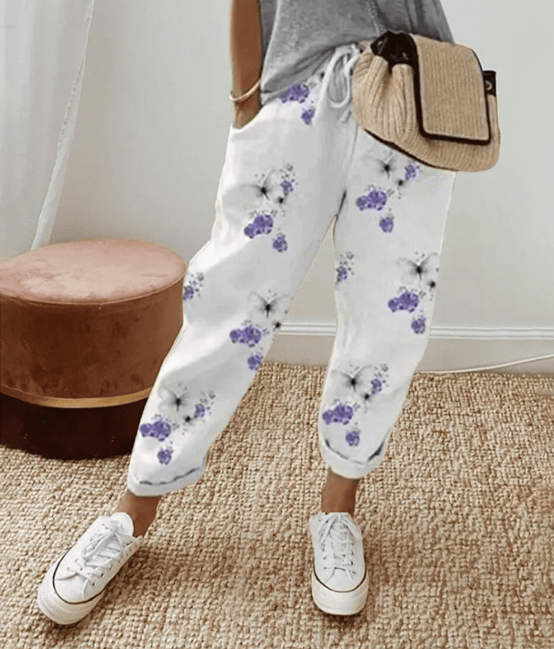 Pantalon blanc à motifs floraux violets, porté avec un t-shirt gris et un sac en osier. Style décontracté, mode féminine tendance.