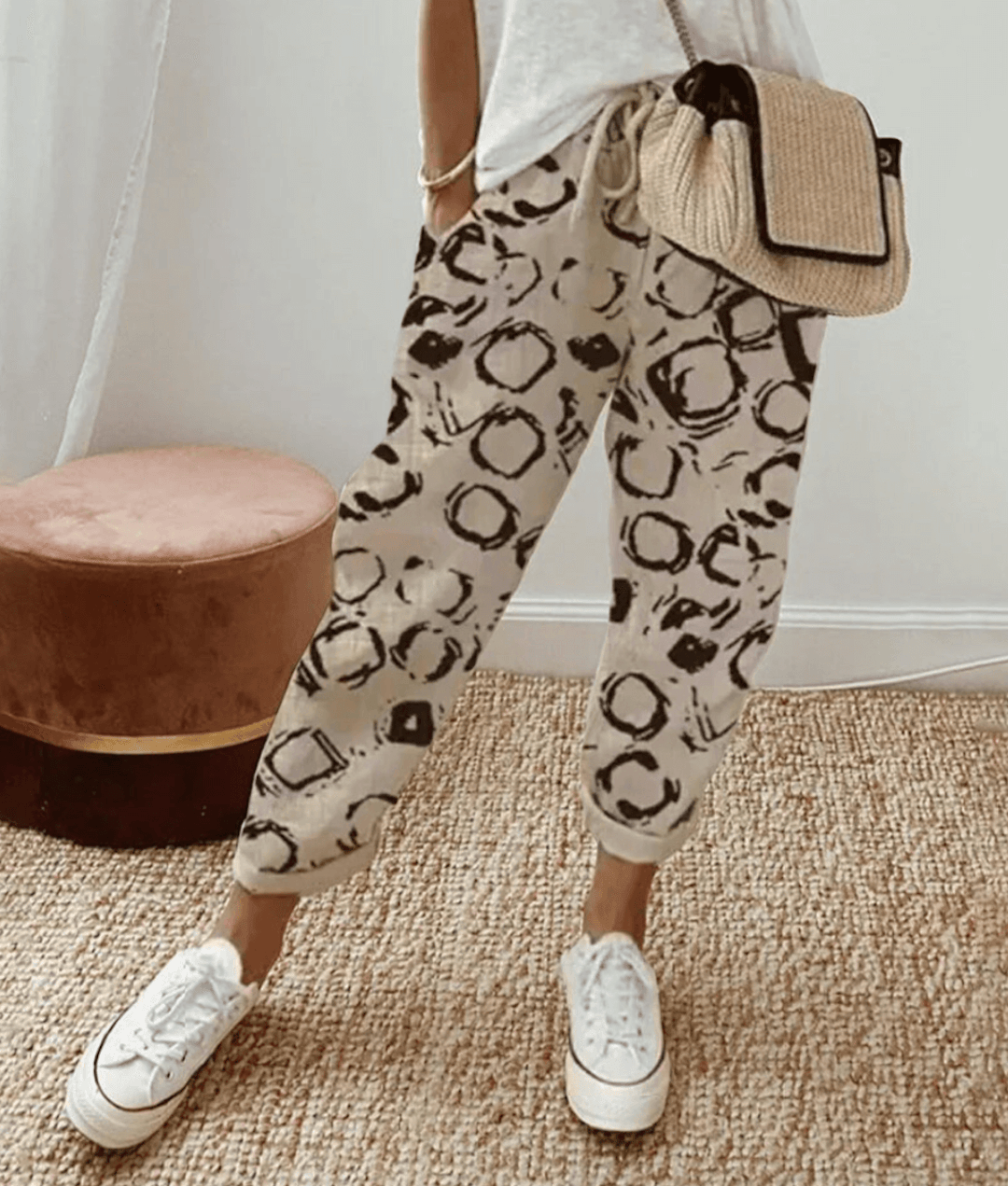 Pantalon beige à motifs noirs abstraits, porté avec un t-shirt blanc et un sac en paille. Style décontracté, chaussures blanches, intérieur moderne.