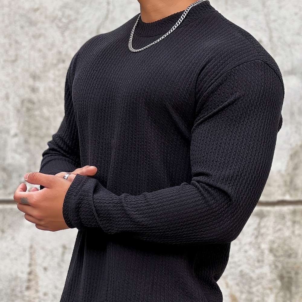 Homme en pull noir texturé, chaîne argentée, posant devant un mur gris. Mode masculine, style décontracté, accessoires tendance.