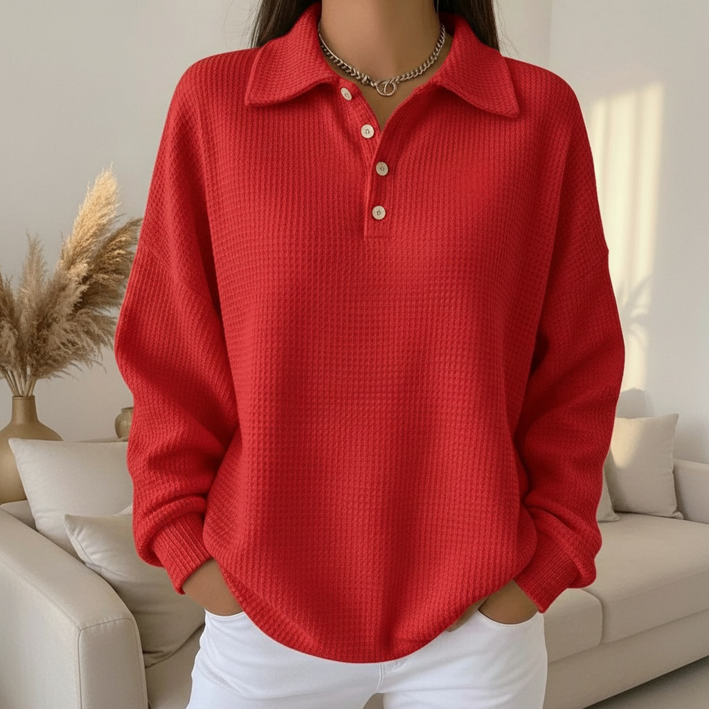 Femme portant un pull rouge à col polo et boutons, posant dans un salon moderne avec canapé beige et plante décorative. Mode automne-hiver tendance.