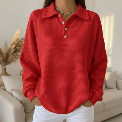 Femme portant un pull rouge à col polo et boutons, posant dans un salon moderne avec canapé beige et plante décorative. Mode automne-hiver tendance.