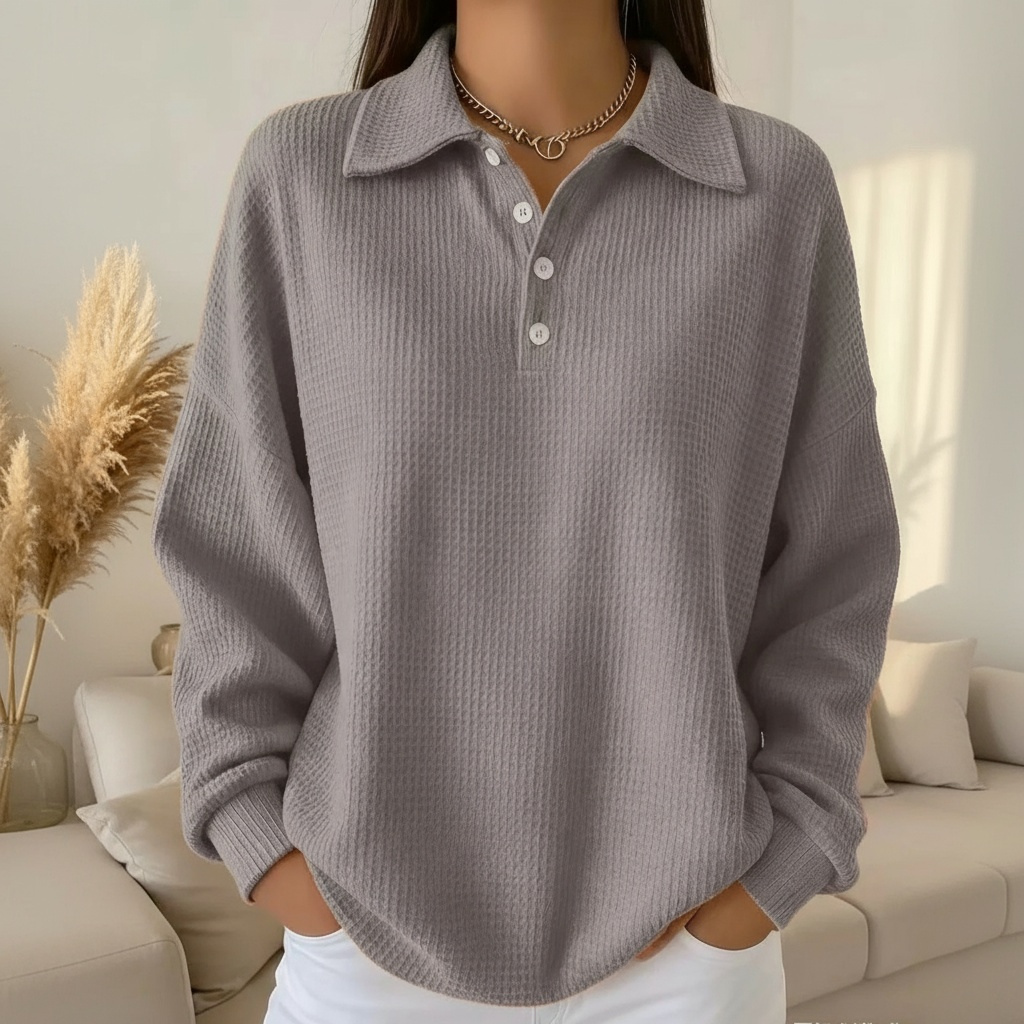 Femme portant un pull gris à col polo, manches longues, dans un salon moderne avec canapé beige et plantes décoratives. Mode automne-hiver élégante.
