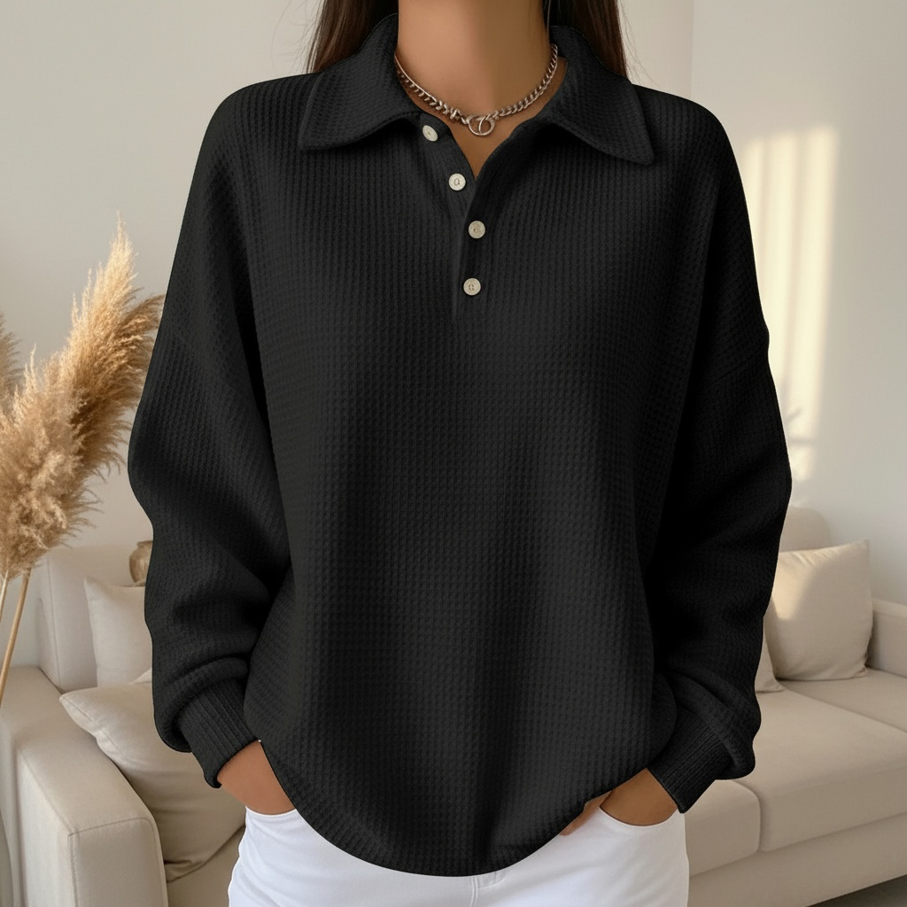 Femme portant un pull noir texturé avec col polo et boutons, posant dans un salon moderne. Mode automne, style décontracté, élégance minimaliste.