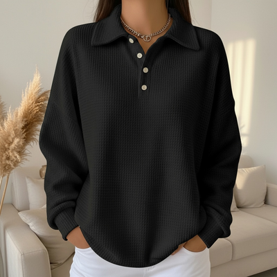 Femme portant un pull noir texturé avec col polo et boutons, posant dans un salon moderne. Mode automne, style décontracté, élégance minimaliste.