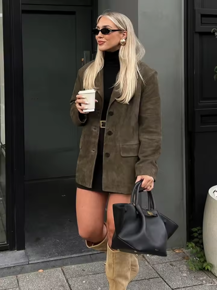 Femme élégante en manteau marron, robe noire, bottes beiges, lunettes de soleil, tenant un café et un sac noir, devant une porte moderne.