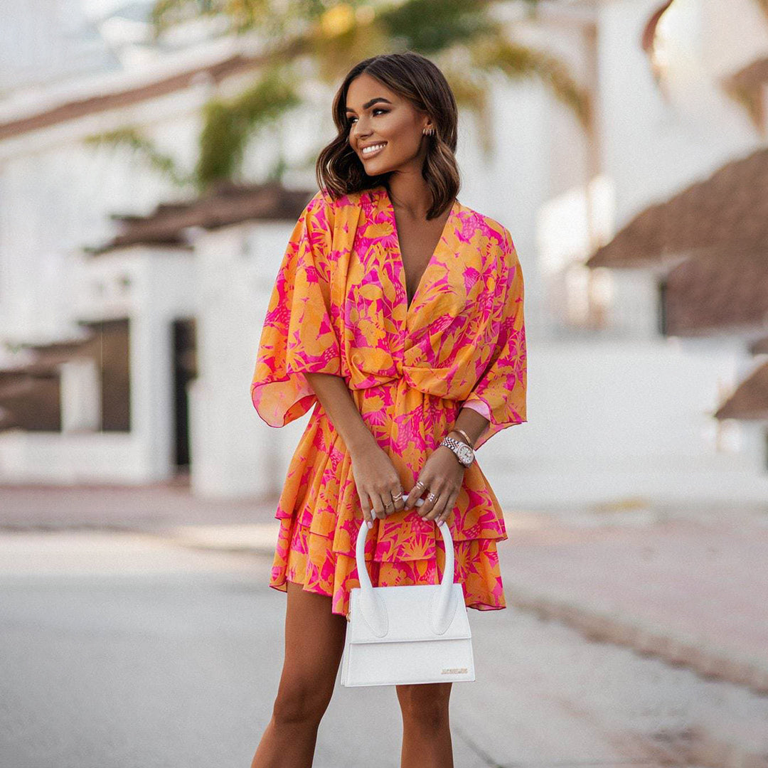Robe d'été rose et orange à motifs tropicaux, femme souriante tenant un sac blanc, fond urbain flou, mode féminine élégante et colorée.