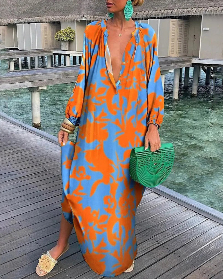 Robe longue orange et bleue à motifs floraux, portée par une femme sur un ponton en bois, avec sac à main vert et boucles d'oreilles assorties.