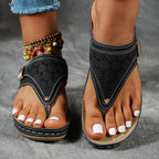 Sandales noires en cuir avec motifs ajourés portées par une femme, ongles blancs, bracelet de cheville coloré, sur fond gris. Mode estivale élégante.