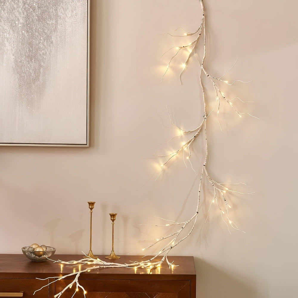 Branche lumineuse décorative avec guirlandes LED, posée sur un meuble en bois, à côté de chandeliers dorés et d'un tableau moderne.