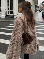 Femme portant un pull en maille épaisse beige et un sac à dos marron, traversant une rue urbaine. Mode automne, style décontracté, look tendance.