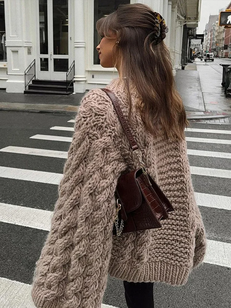Femme portant un pull en maille épaisse beige et un sac à dos marron, traversant une rue urbaine. Mode automne, style décontracté, look tendance.