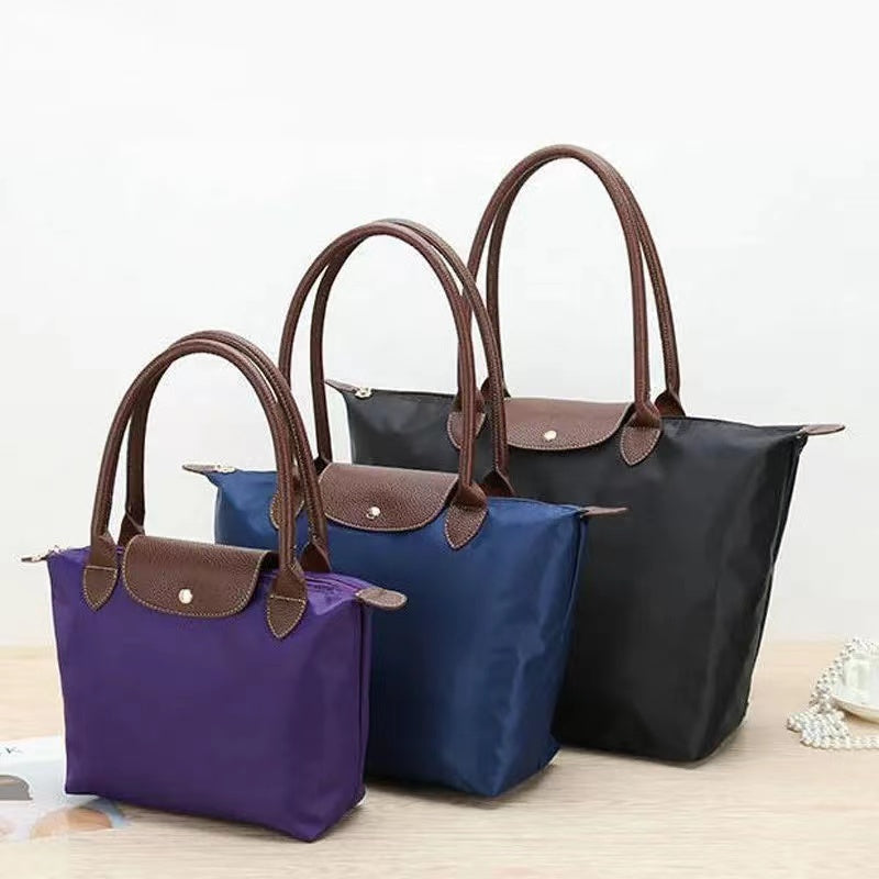 Trois sacs à main en nylon de différentes tailles et couleurs (noir, bleu, violet) avec poignées en cuir marron, posés sur une table en bois.