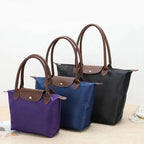 Trois sacs à main en nylon de différentes tailles et couleurs (noir, bleu, violet) avec poignées en cuir marron, posés sur une table en bois.