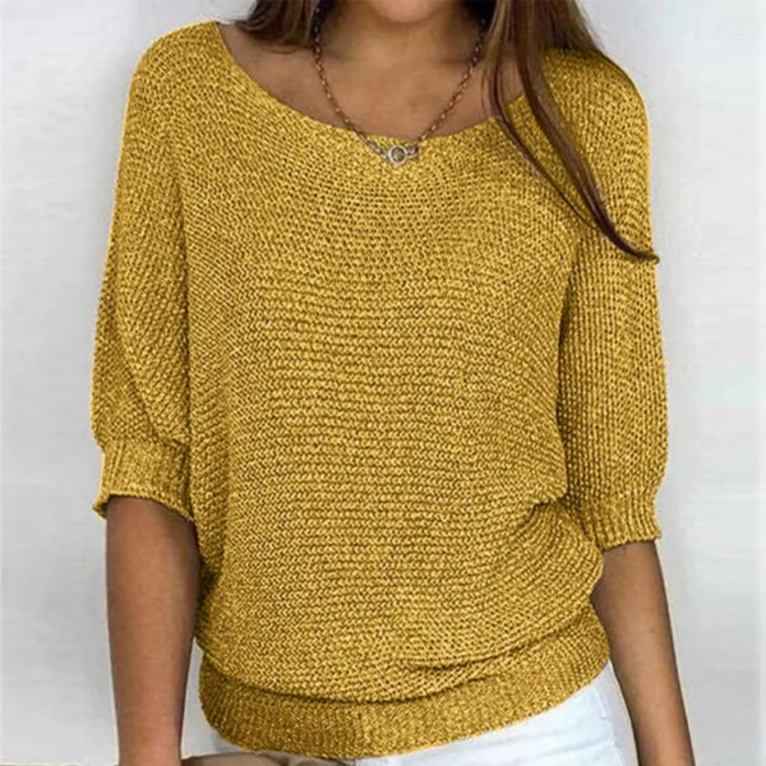 Femme portant un pull jaune moutarde en tricot, manches trois-quarts, col bateau, avec un collier discret, sur fond blanc. Mode automne-hiver tendance.