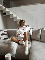 Femme assise sur un canapé gris, portant un pull blanc à motifs de cœurs rouges, chaussettes blanches, avec une tasse de guimauves au premier plan.
