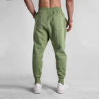 Homme portant un pantalon de jogging vert olive, poches arrière visibles, baskets blanches, sur fond gris. Mode décontractée, vêtements de sport.