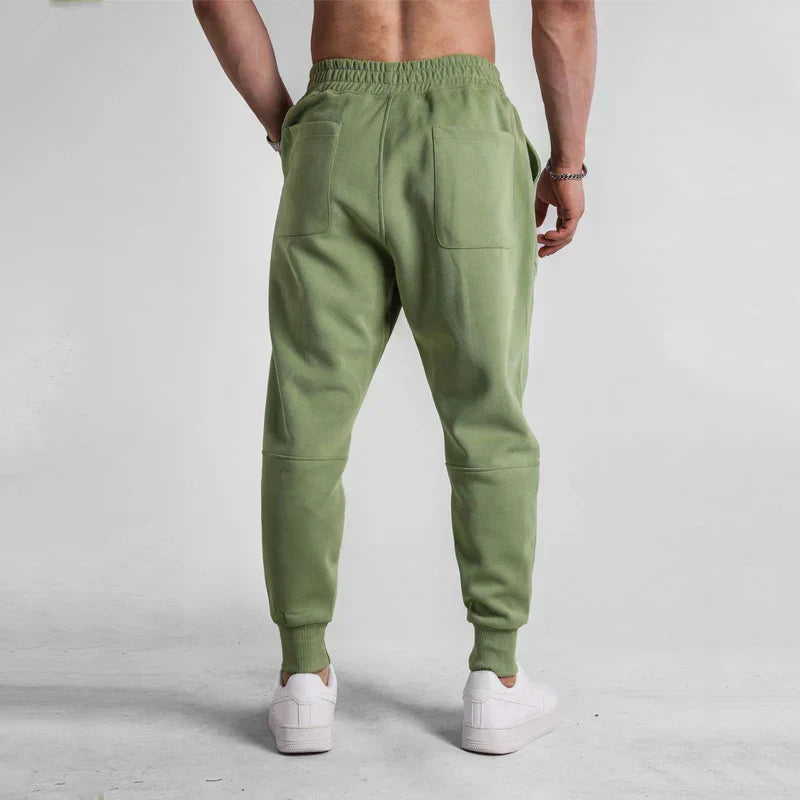 Homme portant un pantalon de jogging vert olive, poches arrière visibles, baskets blanches, sur fond gris. Mode décontractée, vêtements de sport.