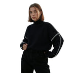 Jeune femme portant un pull noir à col roulé avec détails blancs, posant sur fond blanc. Mode automne-hiver, style décontracté et élégant.