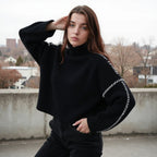 Jeune femme en pull noir à col roulé, posant sur un toit urbain. Mode automne-hiver, style décontracté, paysage urbain en arrière-plan.