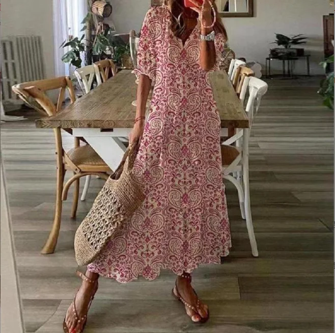 Robe longue bohème rose à motifs floraux, portée par une femme avec un sac en osier, dans une salle à manger moderne avec table en bois et chaises.