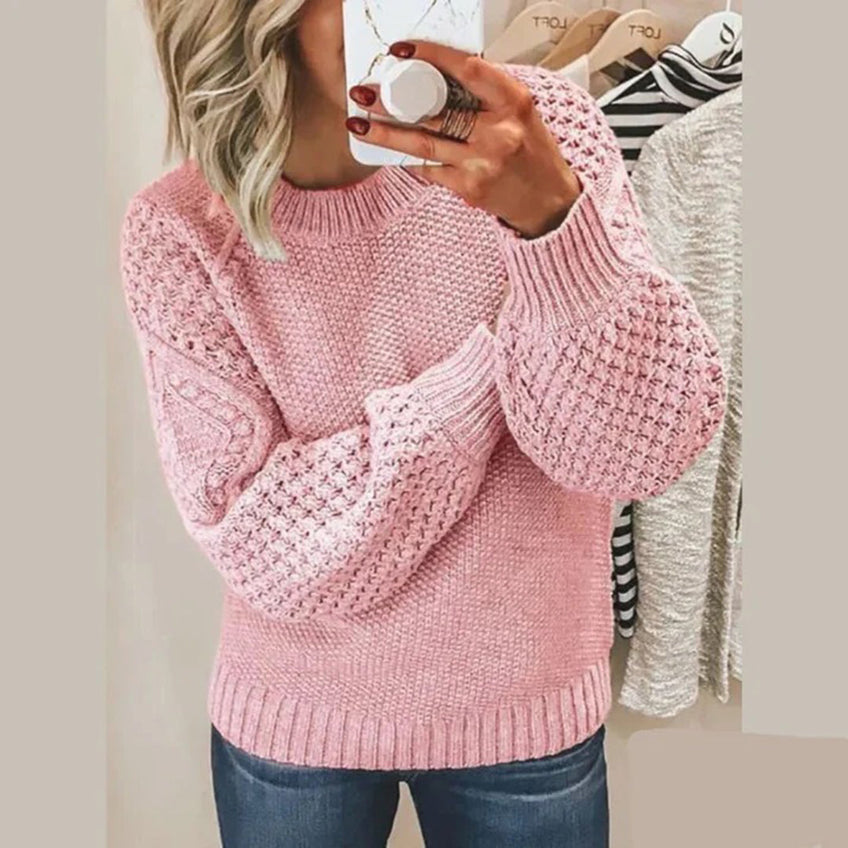 Femme portant un pull rose en tricot texturé, manches longues, tenant un smartphone devant un miroir. Mode automne-hiver, style décontracté.