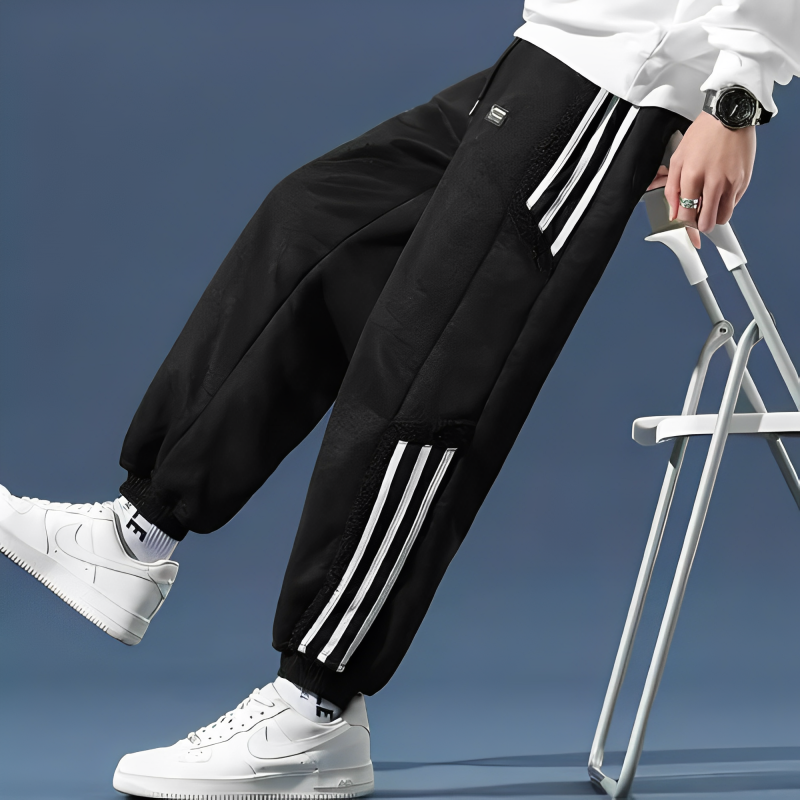 Pantalon de jogging noir avec bandes blanches, porté avec des baskets blanches et une montre noire, sur fond bleu. Mode urbaine tendance.