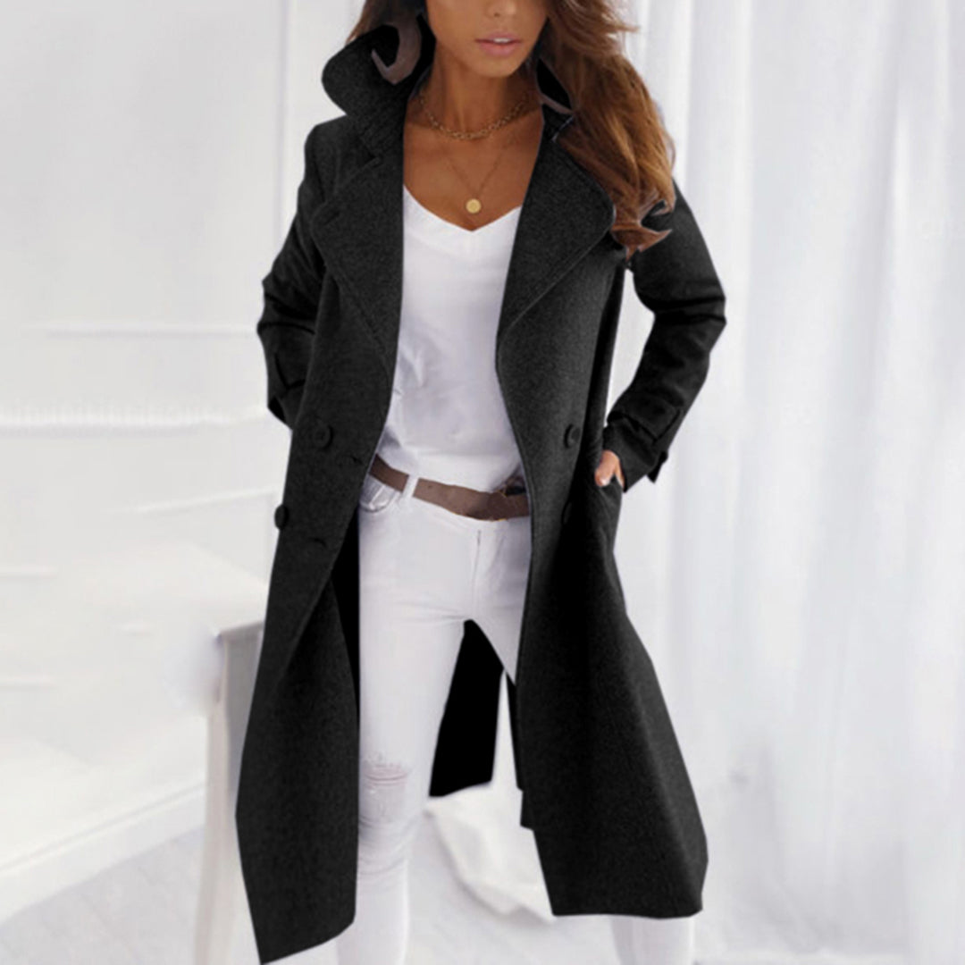 Femme portant un manteau long noir élégant, chemise blanche et pantalon blanc, posant dans un intérieur moderne. Mode automne-hiver chic.