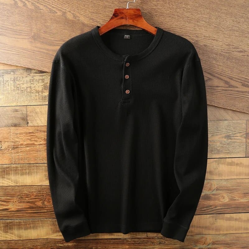 Chemise henley noire à manches longues en coton, avec boutons en bois, suspendue sur un cintre en bois, fond en bois texturé.