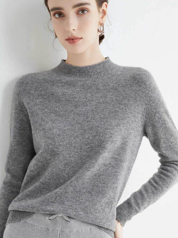 Jeune femme portant un pull gris en cachemire, posant contre un mur blanc. Mode automne-hiver, style élégant et minimaliste.