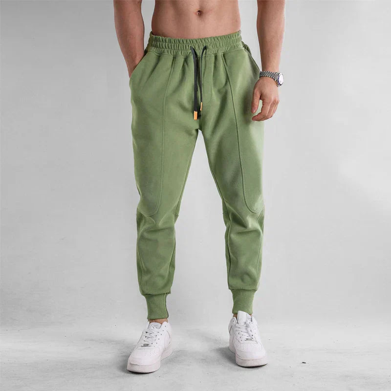 Homme portant un pantalon de jogging vert avec cordon, poches latérales, montre argentée et baskets blanches. Mode décontractée masculine.