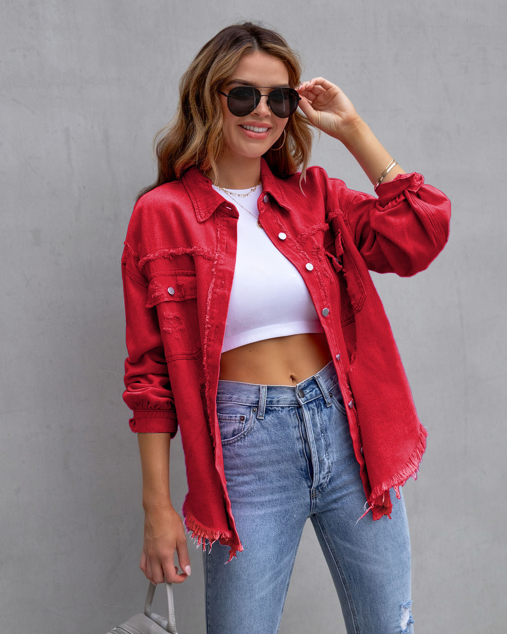 Femme souriante portant une chemise rouge en jean déchiré, un top blanc court et un jean bleu, posant avec des lunettes de soleil devant un mur gris.