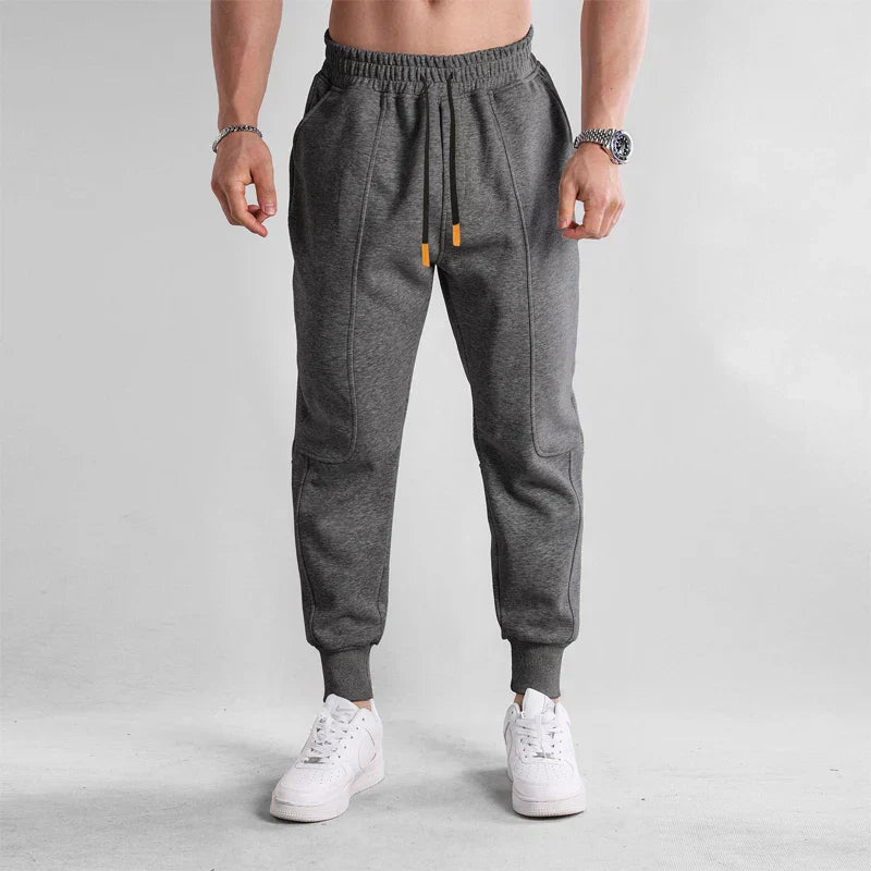 Homme portant un pantalon de jogging gris, taille élastique, avec baskets blanches. Mode décontractée, tenue sportive, style moderne.