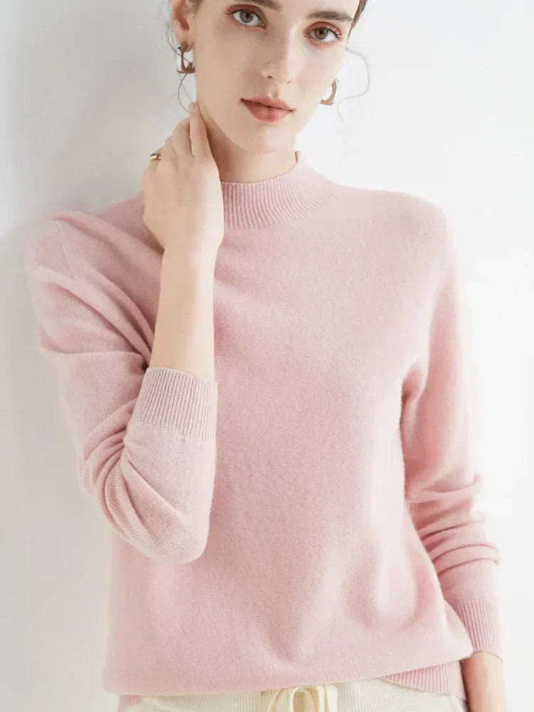 Femme portant un pull rose en cachemire, manches longues, col rond, posant contre un mur blanc. Mode féminine élégante et confortable.