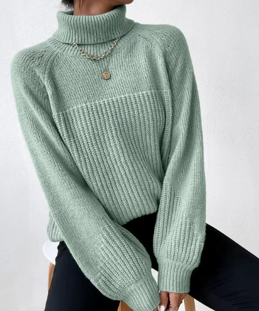 Femme portant un pull vert menthe à col roulé, manches longues, assise sur un tabouret, collier doré, style décontracté et élégant.