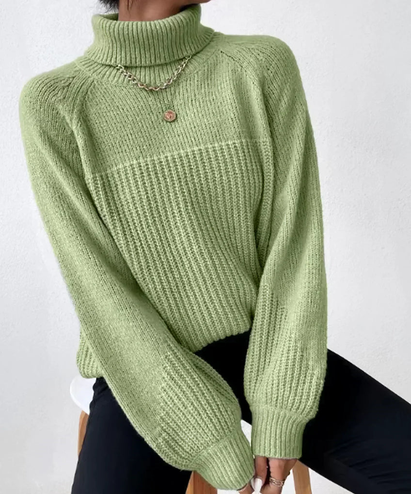 Pull vert en maille côtelée avec col roulé, manches longues, porté par une personne assise. Style décontracté, tendance automne-hiver.