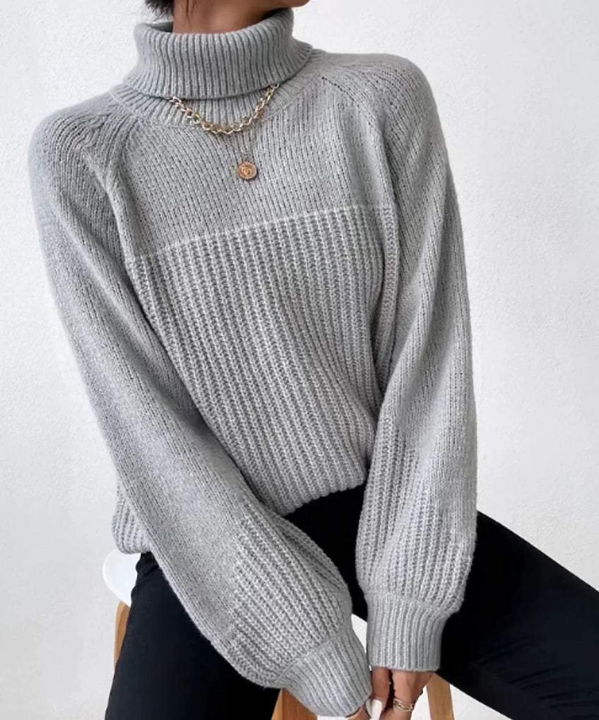 Femme portant un pull gris à col roulé en tricot, manches longues, avec un collier doré, assise sur un tabouret. Mode hiver élégante et confortable.