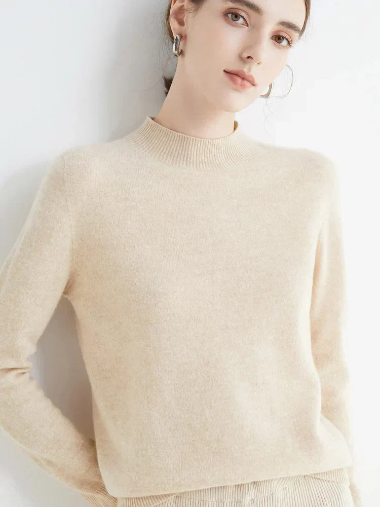 Femme portant un pull en cachemire beige, col rond, manches longues, posant contre un mur blanc. Mode élégante, tendance automne-hiver.